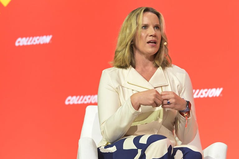 Image of a person (Catharina Lavers Mallet, former VP and general manager for Maxis and The Sims (Electronic Arts)), speaking on stage at Collision 2023. Catharina is wearing a headset mic and gesturing with both hands. Catharina appears to be speaking. Catharina is sitting on a chair. Screens behind the former VP bear the Collision logo.