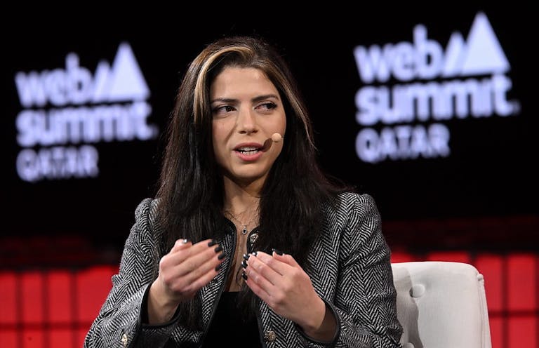 A person (astronaut and entrepreneur Sara Sabry) sitting in an armchair. Sara is wearing a headset mic and gesturing with both hands. Sara appears to be speaking. Screens behind Sara bear the Web Summit Qatar logo. This is Centre Stage on Day 1 of Web Summit Qatar.