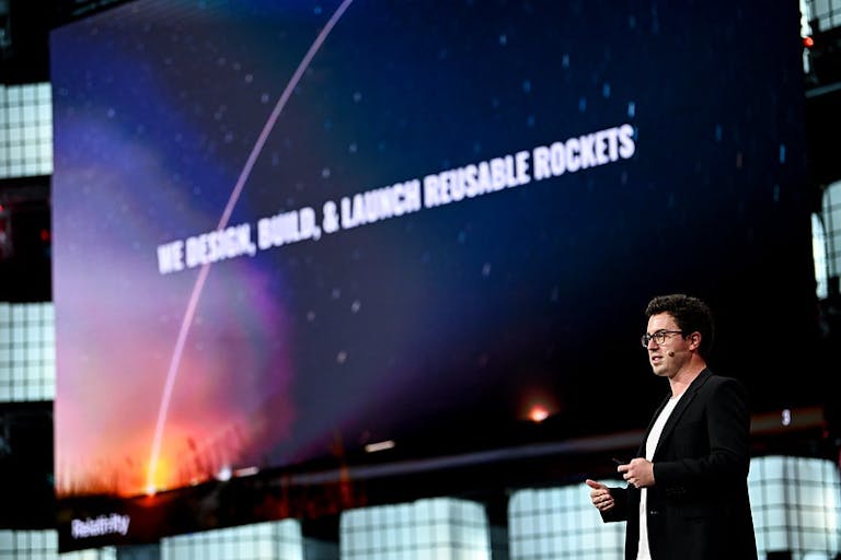 A standing person (Relativity Space co-founder and CEO Tim Ellis) wearing a headset mic and holding a presentation clicker. Behind Tim, a screen bears the message 'We design, build and launch reusable rockets'. This is Centre Stage on Day 3 of Web Summit Qatar.