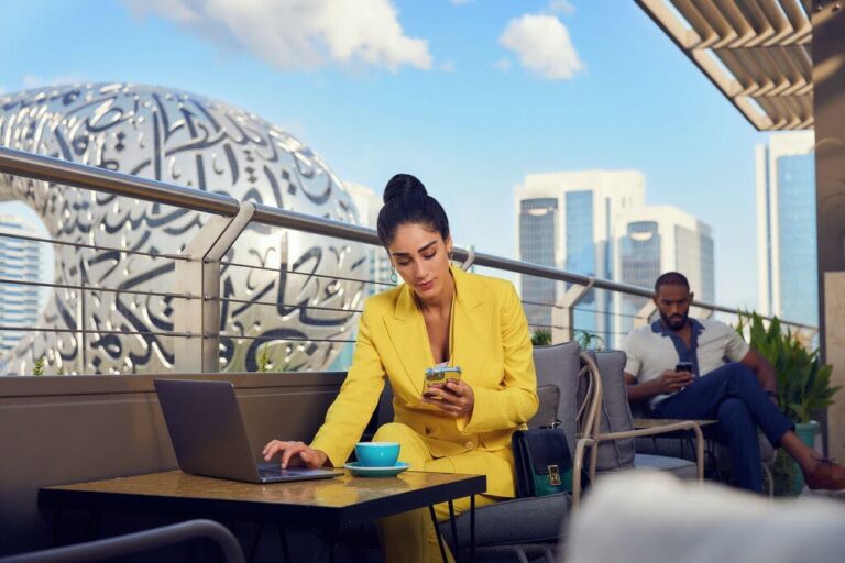 A person sits at a low table on a balcony. They are looking at a smartphone that they're holding in their left hand, and touching the trackpad of a laptop with their right hand. Visible beyond the balcony is the skyline of Dubai, including most prominently a structure that appears to be made of metal, with Arabic calligraphy inlaid in it in glass. This is Dubai's Museum of the Future.