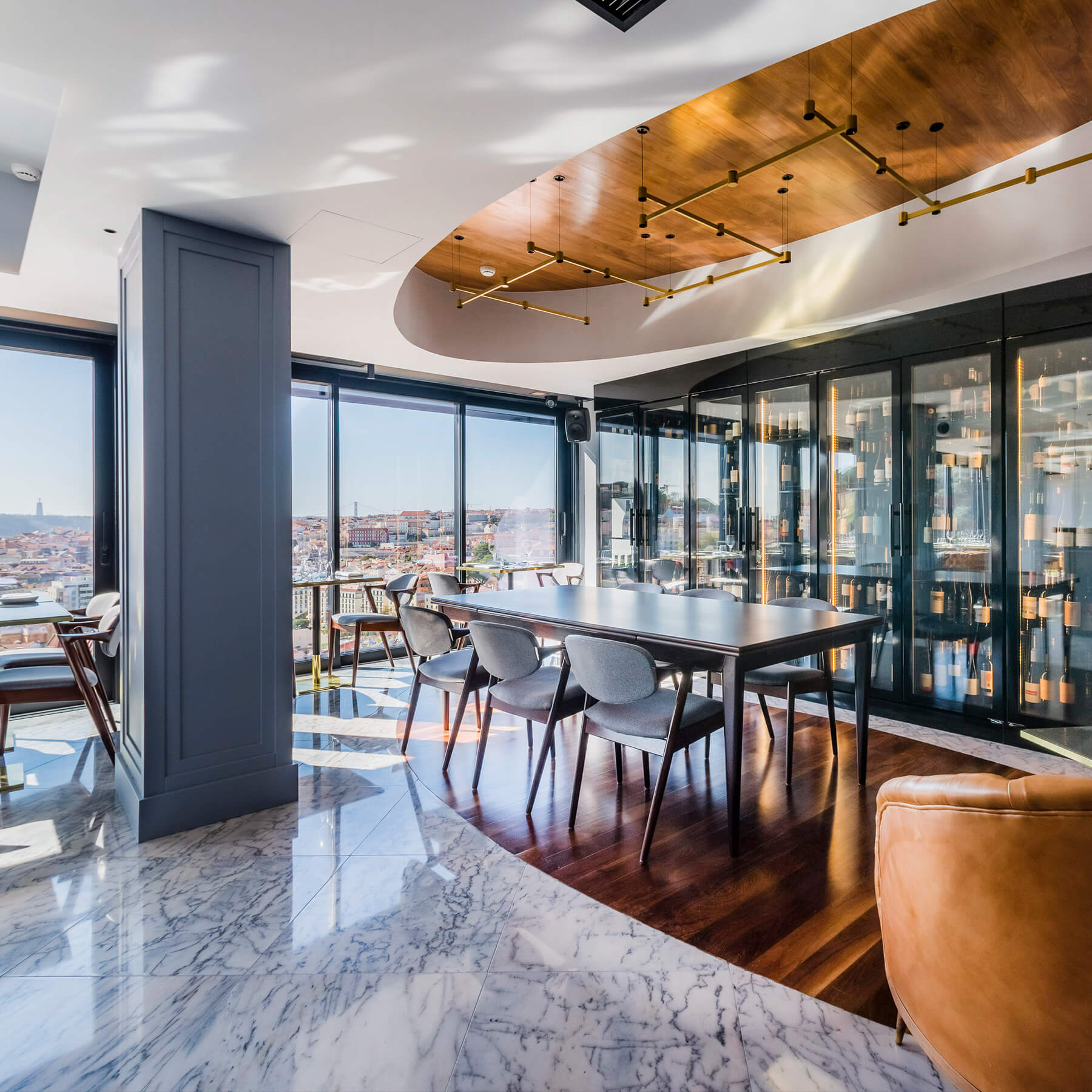 A modern room with marble floors and floor-to-ceiling windows. Tables dot the room, including a table and six chairs that sit atop an area of floorboards in the centre of the space. A recess in the ceiling contains a light fixture consisting of interconnected metal poles. Behind the central table is a bank of glass-fronted cabinets containing bottles of wine.