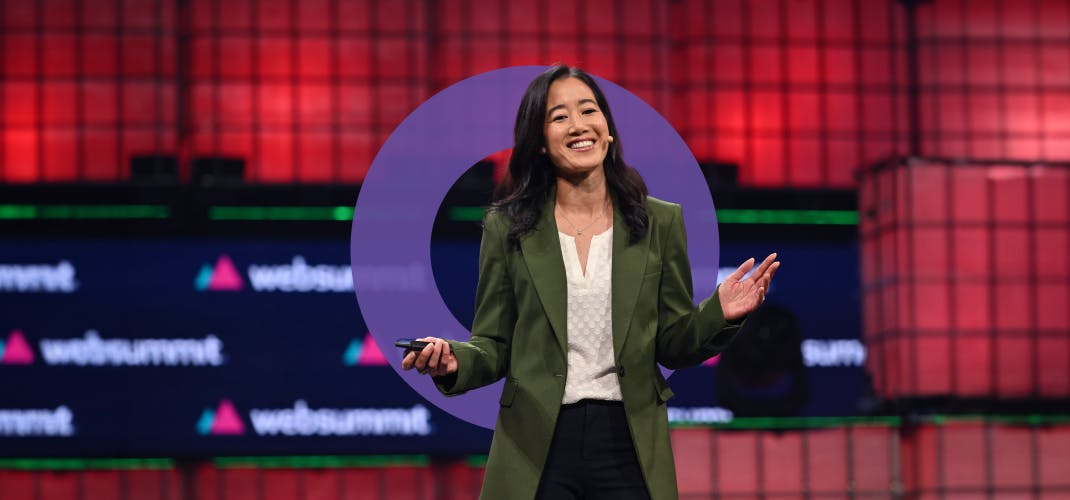 A person stands on stage, pictured from the waist up. They are smiling. They're wearing a headset mic, gesturing with both hands, and holding a presentation clicker in their right hand. The Web Summit logo is visible in several places behind them. This is Centre Stage at Web Summit.