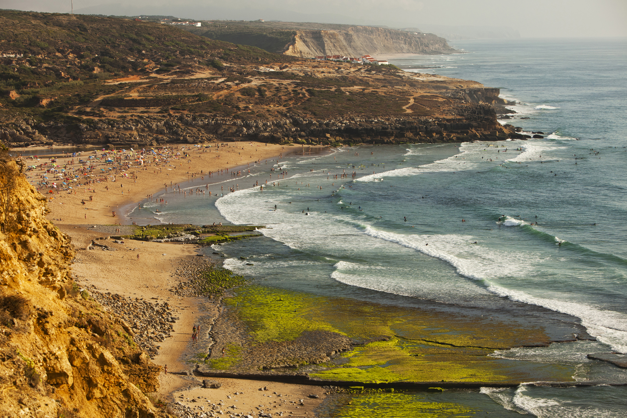 A sandy beach viewed from atop a cliff. People are dotted along the sand and in the water. White foam caps the waves. A rugged coastline disappears into the distance.