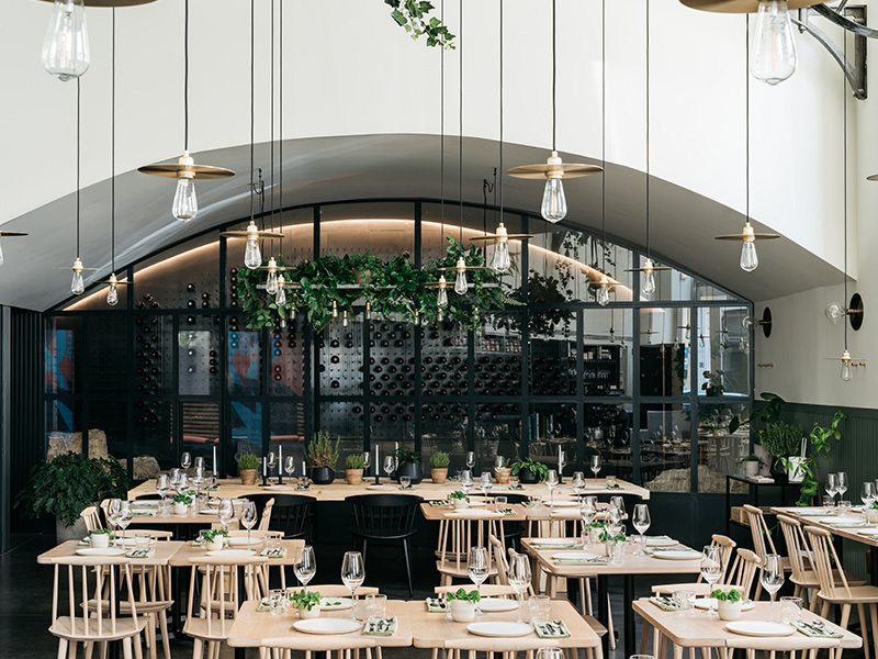 A high-ceilinged room crowded with tables set for dinner with plates, silverware and wine glasses. Lights consisting of metal discs and edison bulbs hang from the ceiling on long cables. Towards the rear of the room is an area below a low vaulted ceiling. Here, a wall of glass separates the rest of the room from what appears to be a wine cellar.