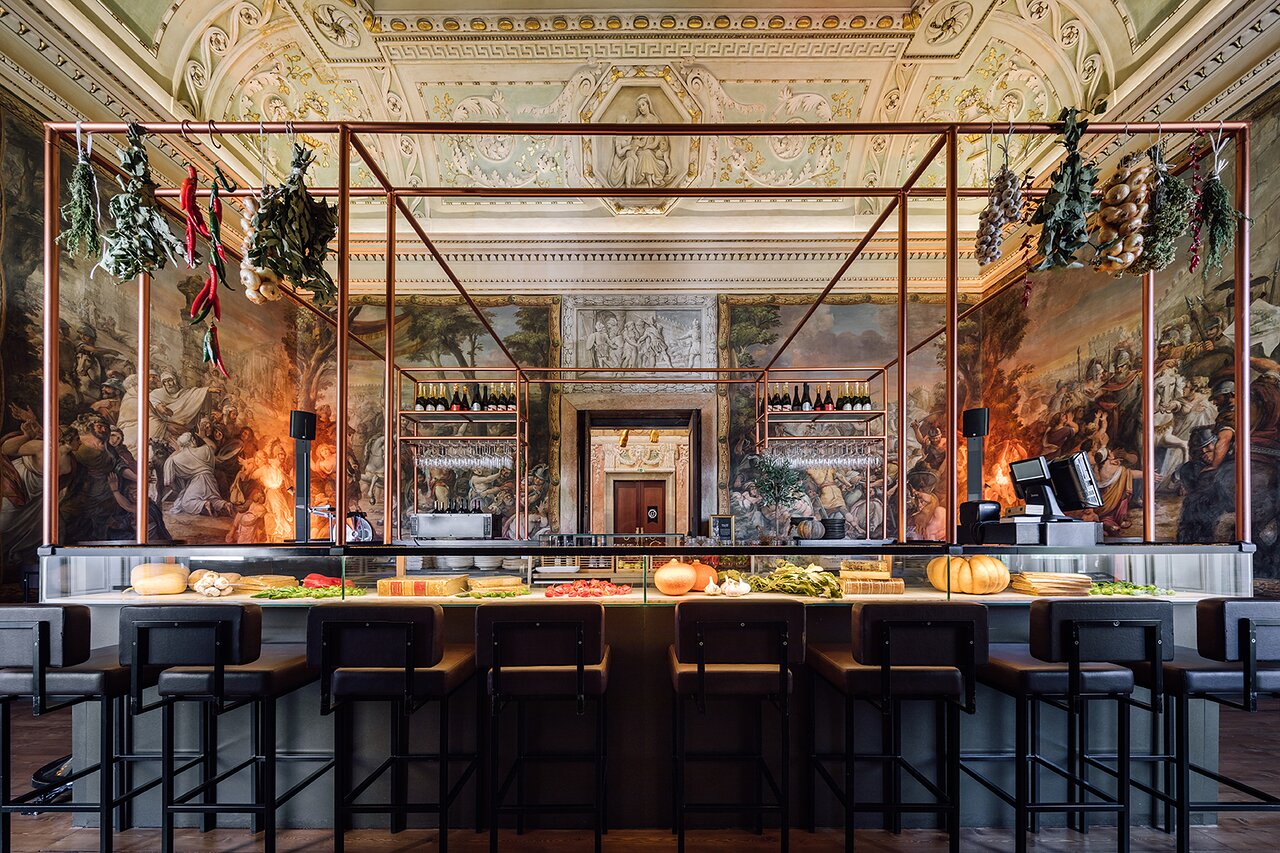 A high-ceilinged room with walls painted floor to ceiling with a Renaissance-style battle scene. There is an elaborately carved vaulted ceiling, featuring a relief of a woman and child. In the centre of the room stands a modern bar, with gourds, vegetables and books displayed under a glass counter. Herbs and spices hang from an industrial-style metal scaffold that incorporates shelves for glasses and bottles. A cash register sits on the right of the bar, and eight high chairs with low backs face the bar.