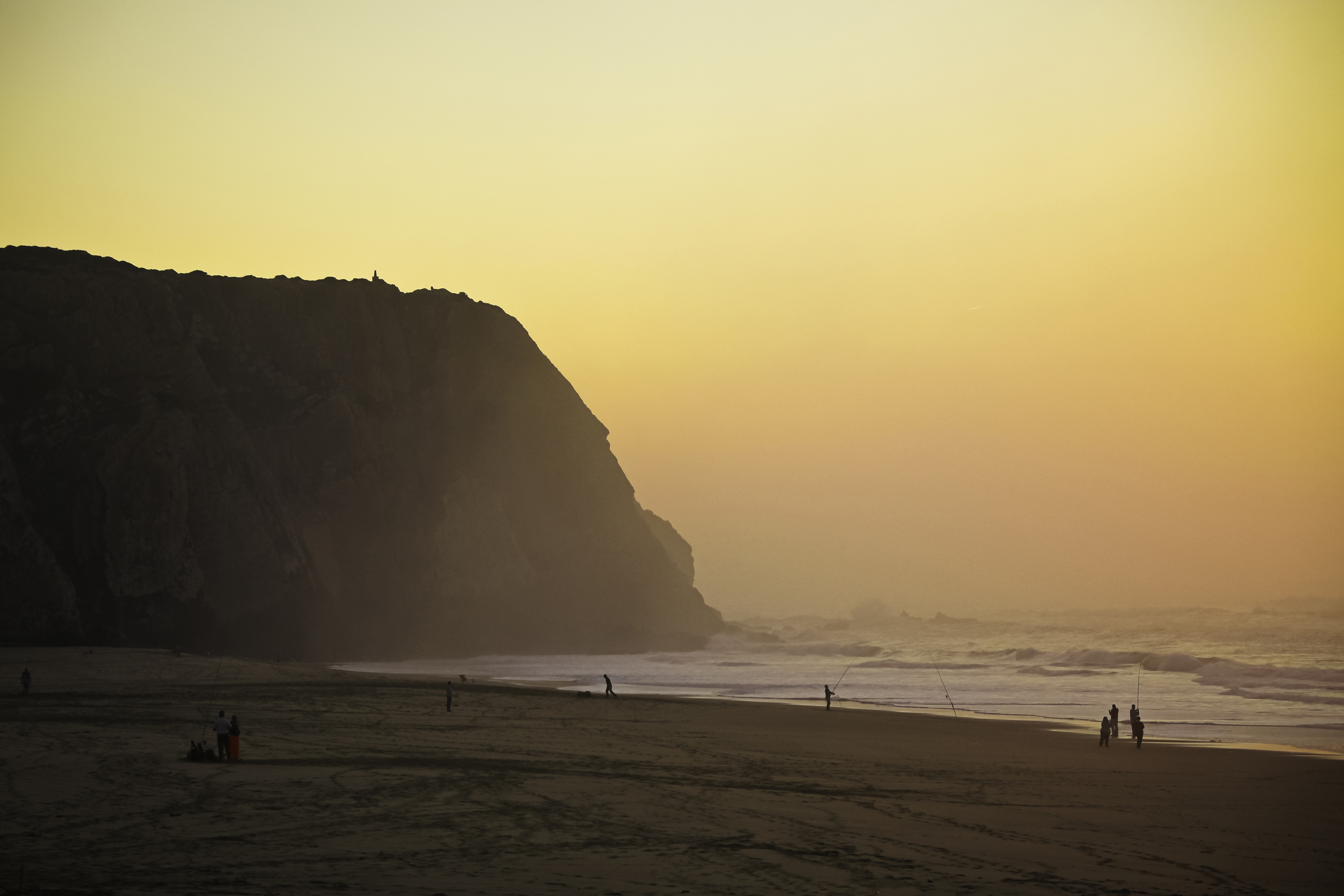 A sandy beach at sunset. A rocky cliff juts into the water. The beach is quiet, but there are several people fishing where the sand meets the sea. The waves are capped with foam.