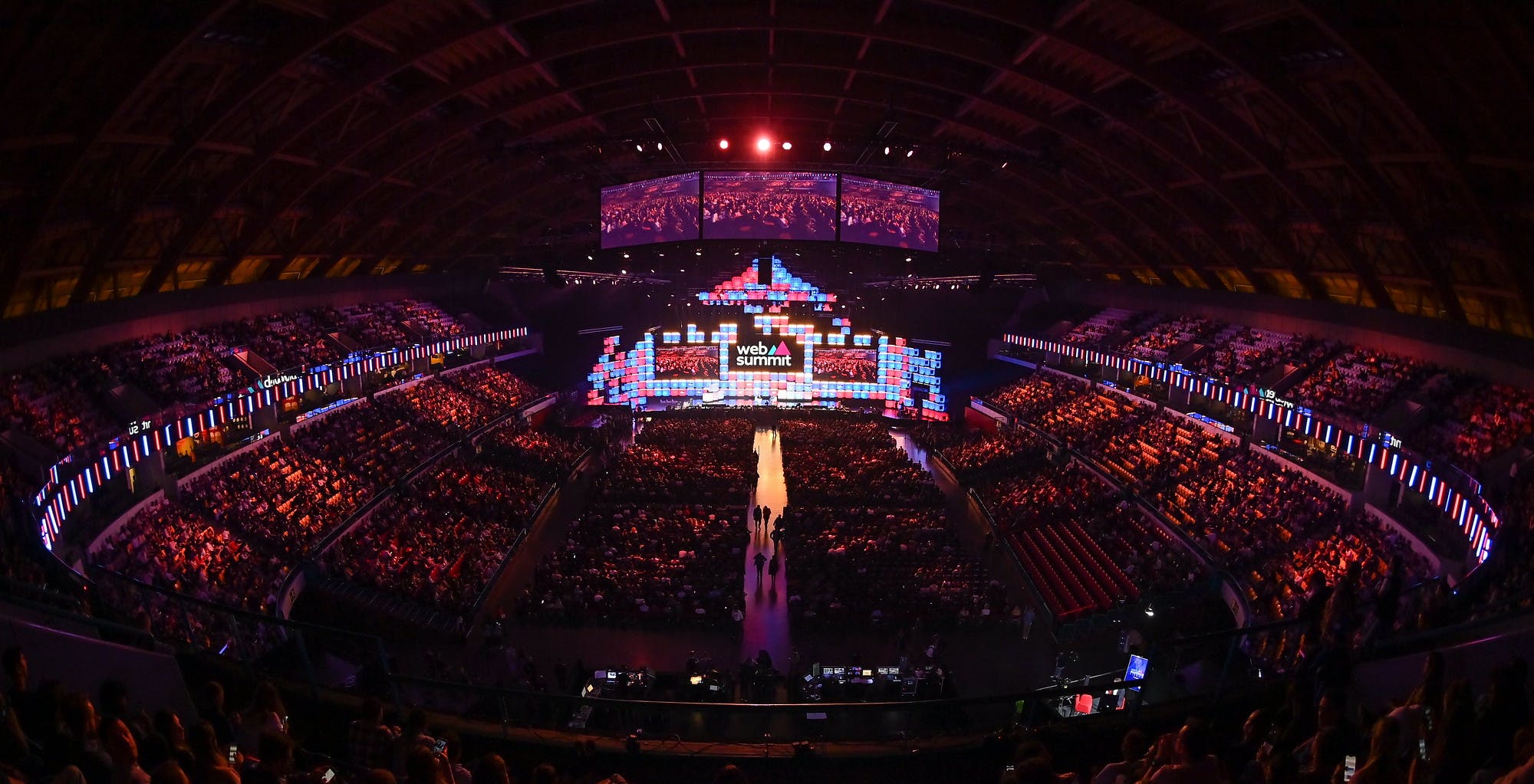 An arena stage viewed from a distance. The stage features the Web Summit logo between two large screens. Three large screens are suspended from the ceiling over a packed crowd. The screens all show shots of the crowd. It is Web Summit Opening Night.